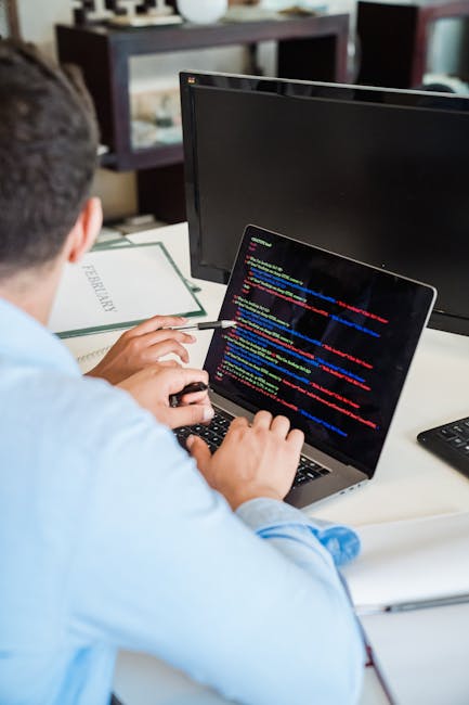A person sitting at a desk working on a laptop with a dark screen displaying lines of colorful computer code. The individual, viewed from behind, is wearing a light blue shirt and is holding a pen in 