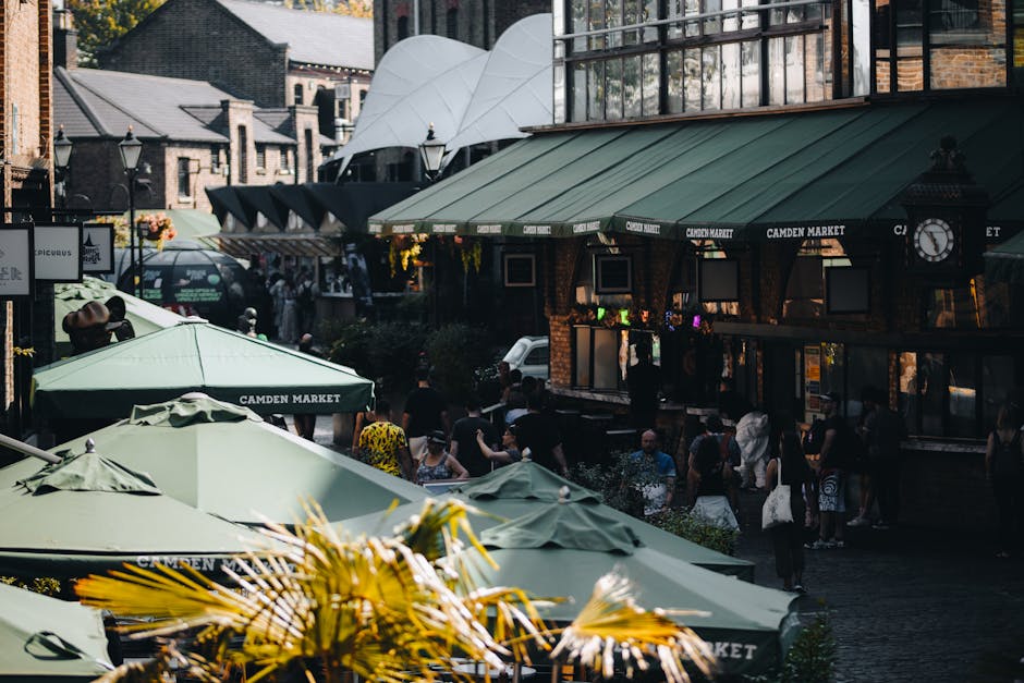 A busy outdoor scene at Camden Market showing a cluster of green and beige umbrellas providing shade over outdoor seating areas. The umbrellas, branded with 'Camden Market,' are positioned in front of