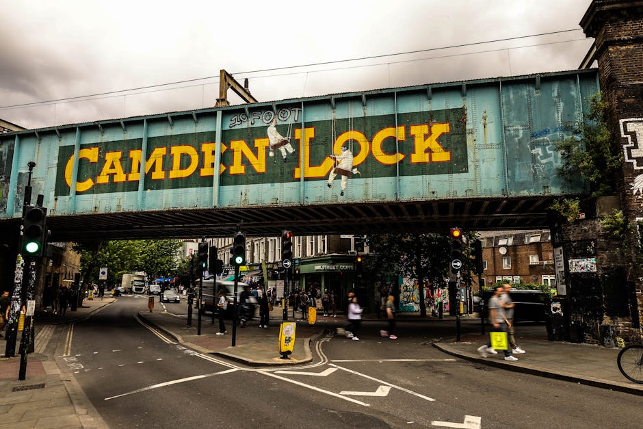 A busy outdoor scene at Camden Market showing a cluster of green and beige umbrellas providing shade over outdoor seating areas. The umbrellas, branded with 'Camden Market,' are positioned in front of